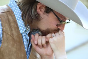 A person in a tan suede vest and polka dot shirt leans forward while bringing their fiance's hand to their mouth.