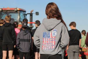 A person wearing a gray hoodie with American flag design stands in a crowd outdoors near a tractor.
