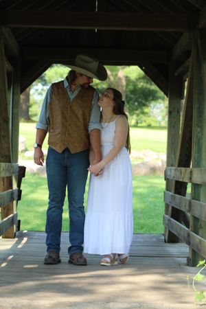 A couple stands together on a covered wooden bridge, with the person in a tan vest and cowboy hat beside someone in white dress.