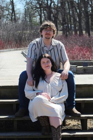Two people sitting together on wooden steps outdoors on a sunny day surrounded by trees and red bushes.