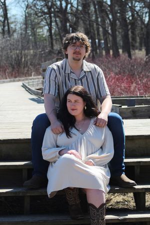 A couple sits together on wooden benches in a park setting with bare trees and red bushes in the background.