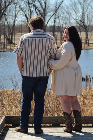 A couple stands on a wooden deck overlooking a lake with bare trees and dried reeds in the background.