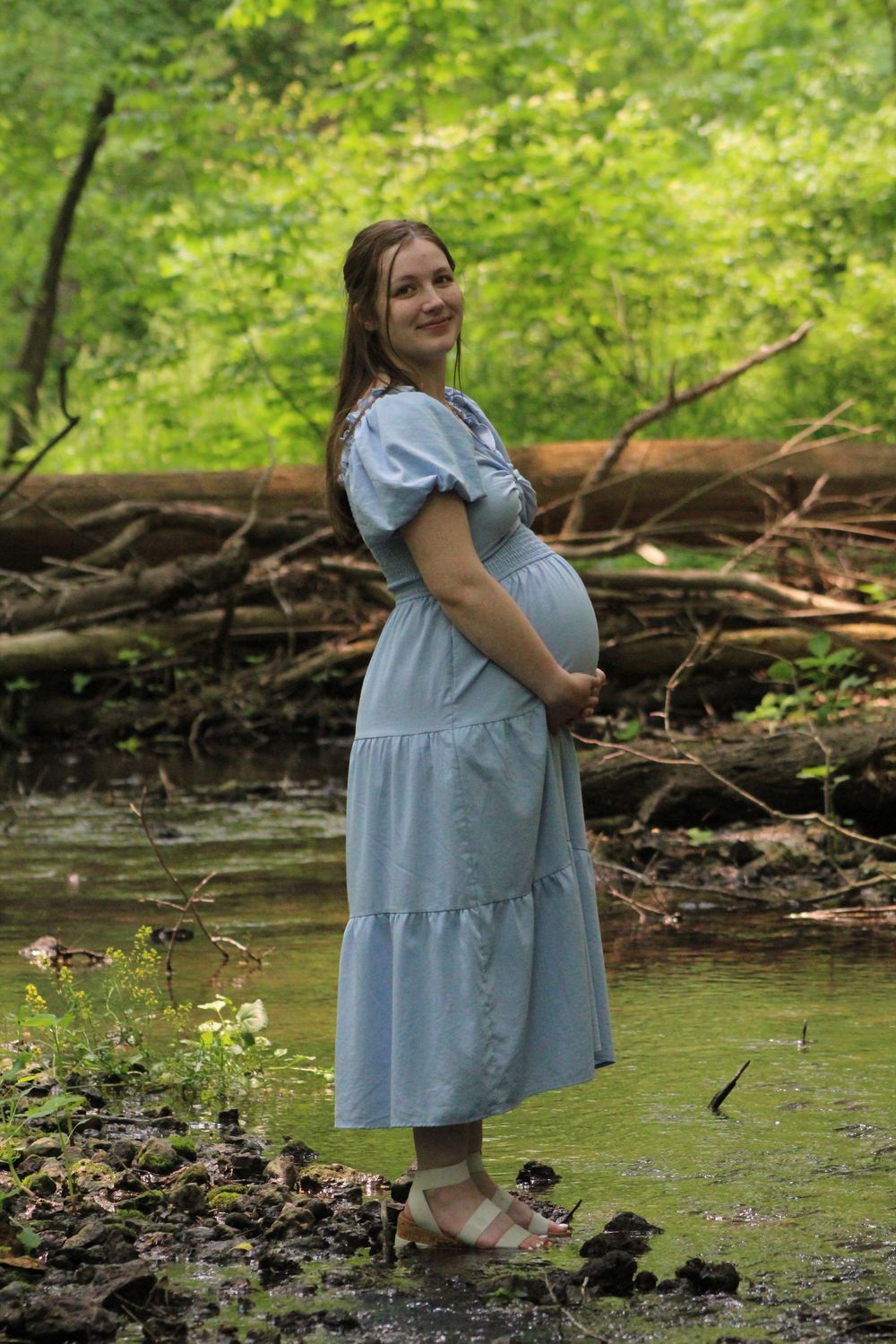 A light blue tiered dress with short puff sleeves is worn in a serene woodland setting by a creek.