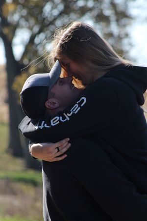Couple embracing and smiling outdoors in autumn, wearing matching black Carhartt hoodies, with trees in the background.