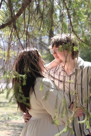 A romantic couple stands beneath drooping willow branches on a sunny spring day in a natural outdoor setting.