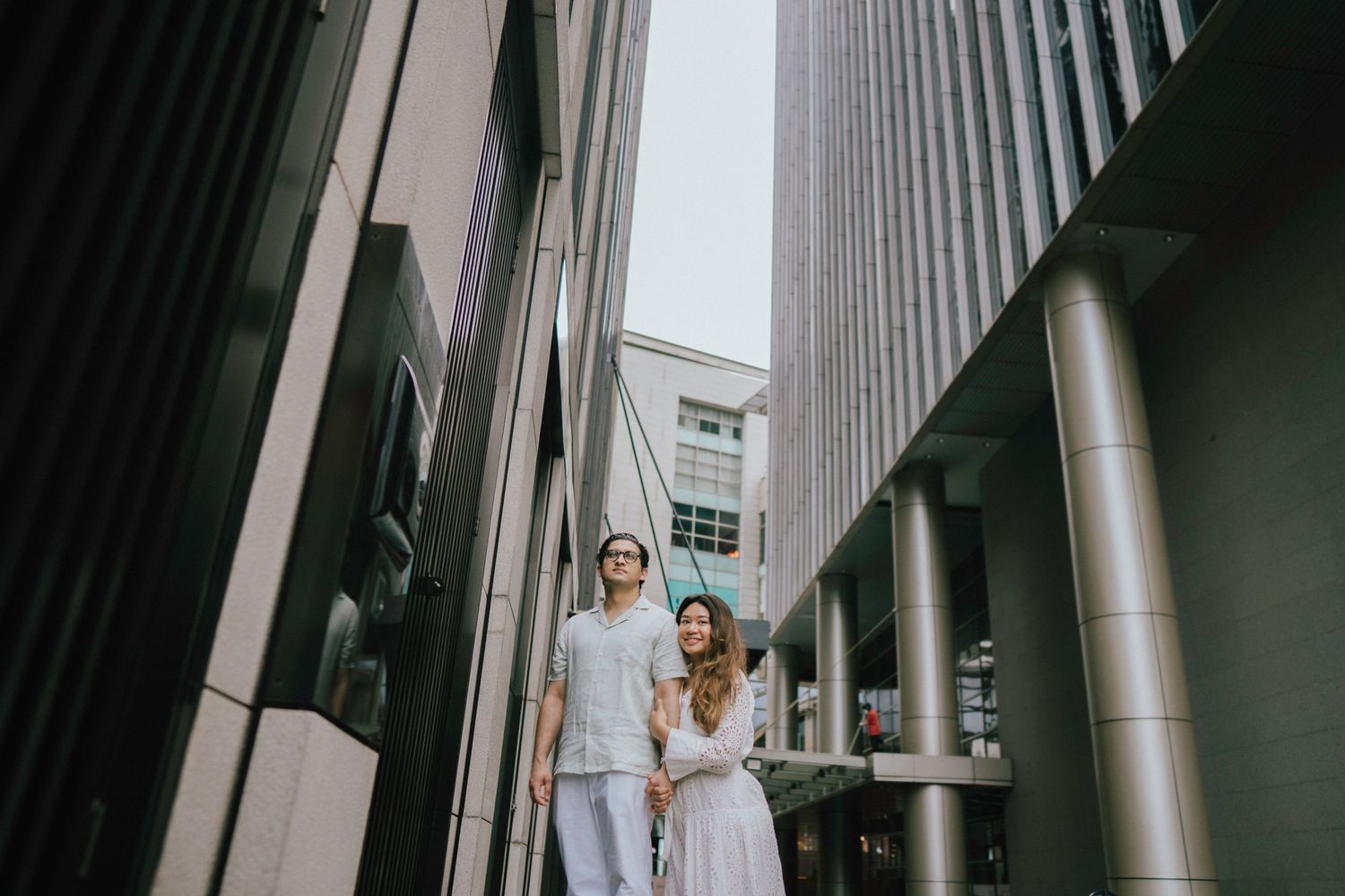 Couple in white casual outfits walking between modern urban buildings during engagement photoshoot.