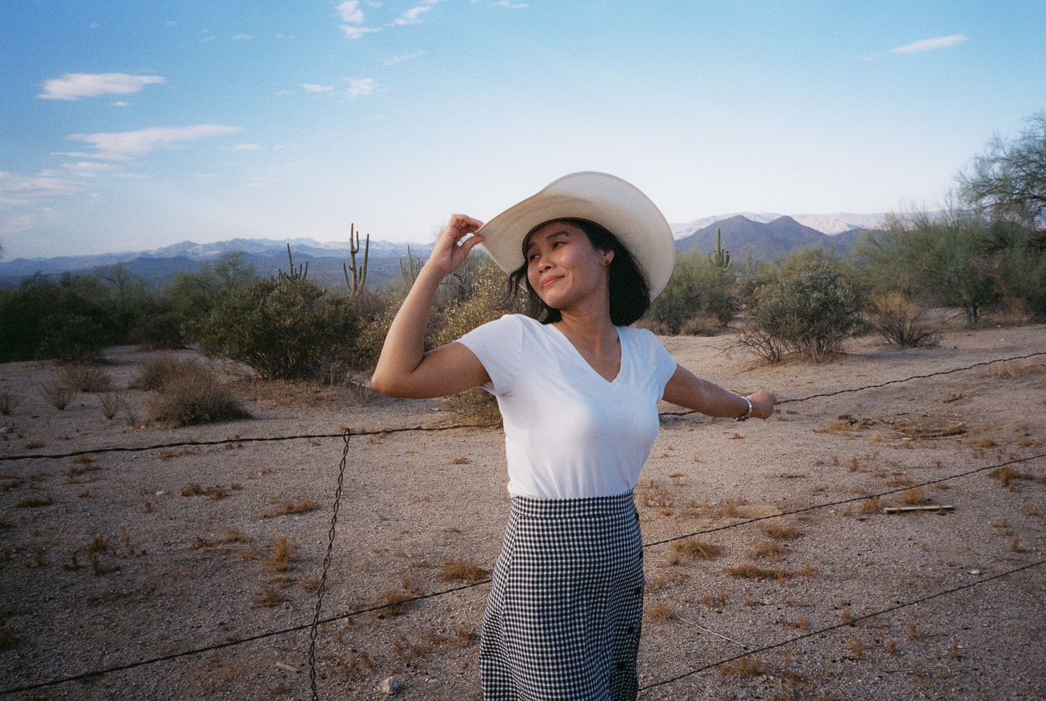 A figure in a white cowboy hat and skirt poses dramatically in a desert landscape with cacti.