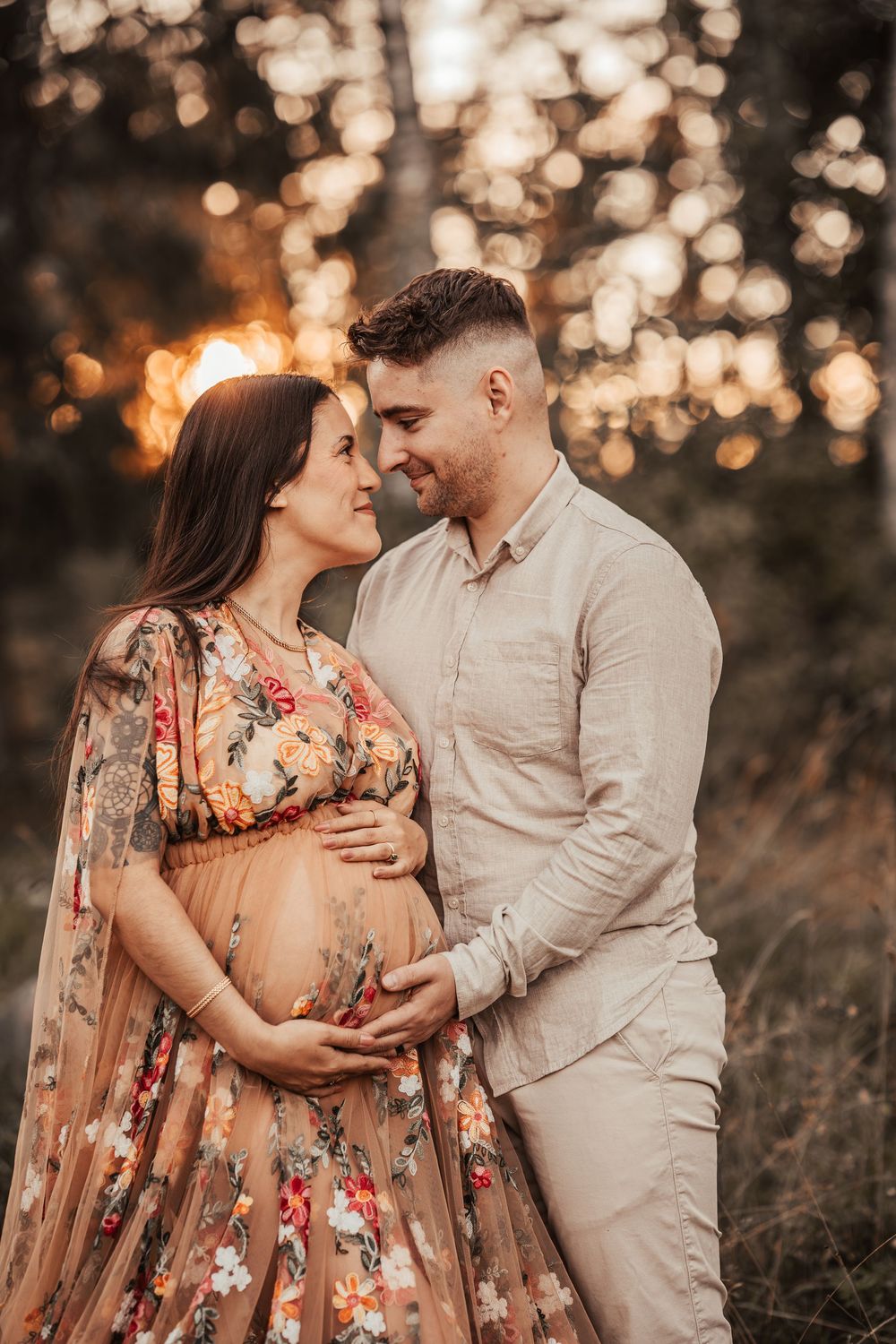 A couple shares an intimate moment at sunset in a field with golden bokeh lighting and the woman wearing a floral dress.