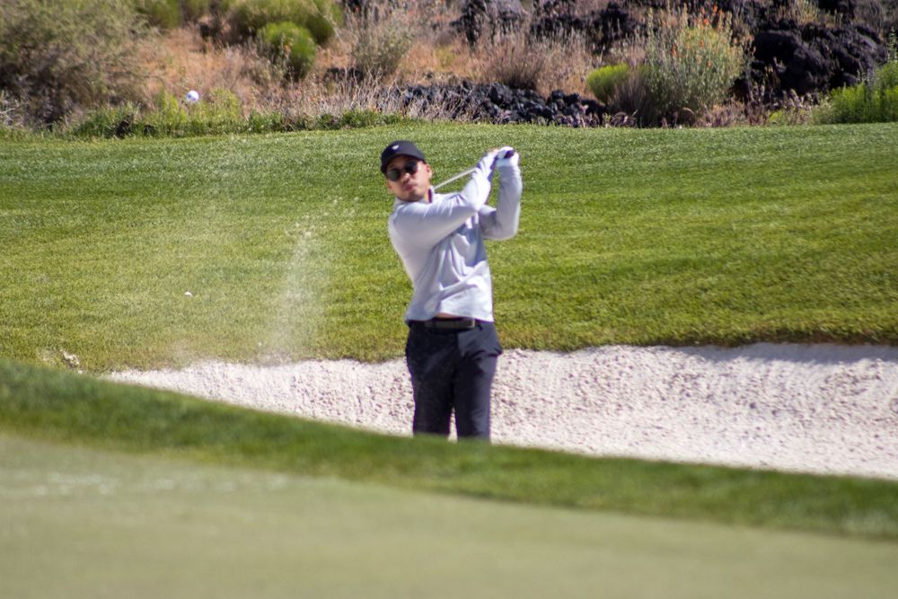Golfer hits shot from sand bunker on desert golf course surrounded by rocky terrain.