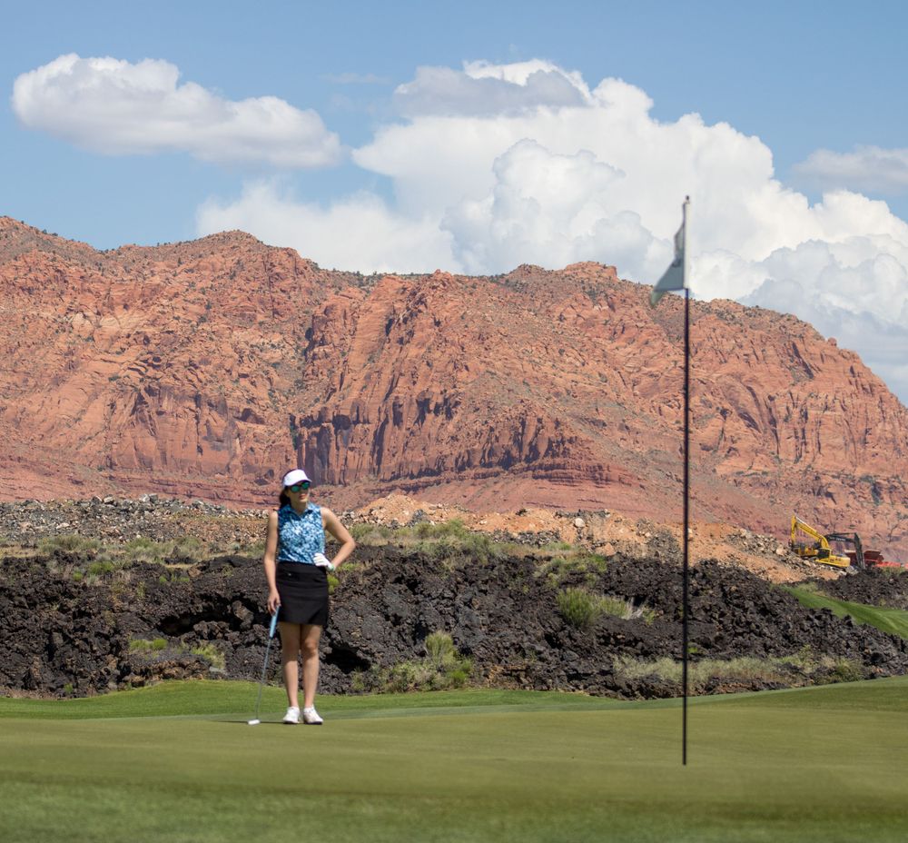 Woman golfer stands near flag on lush green course with stunning red rock mountain backdrop.