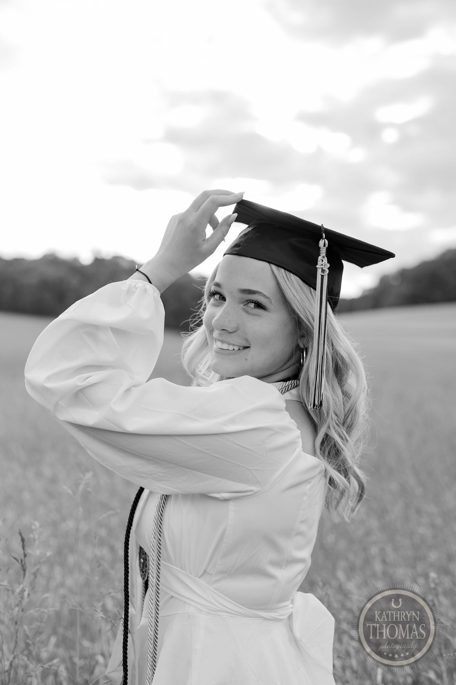 Black and white portrait of graduate adjusting cap in rural field setting.