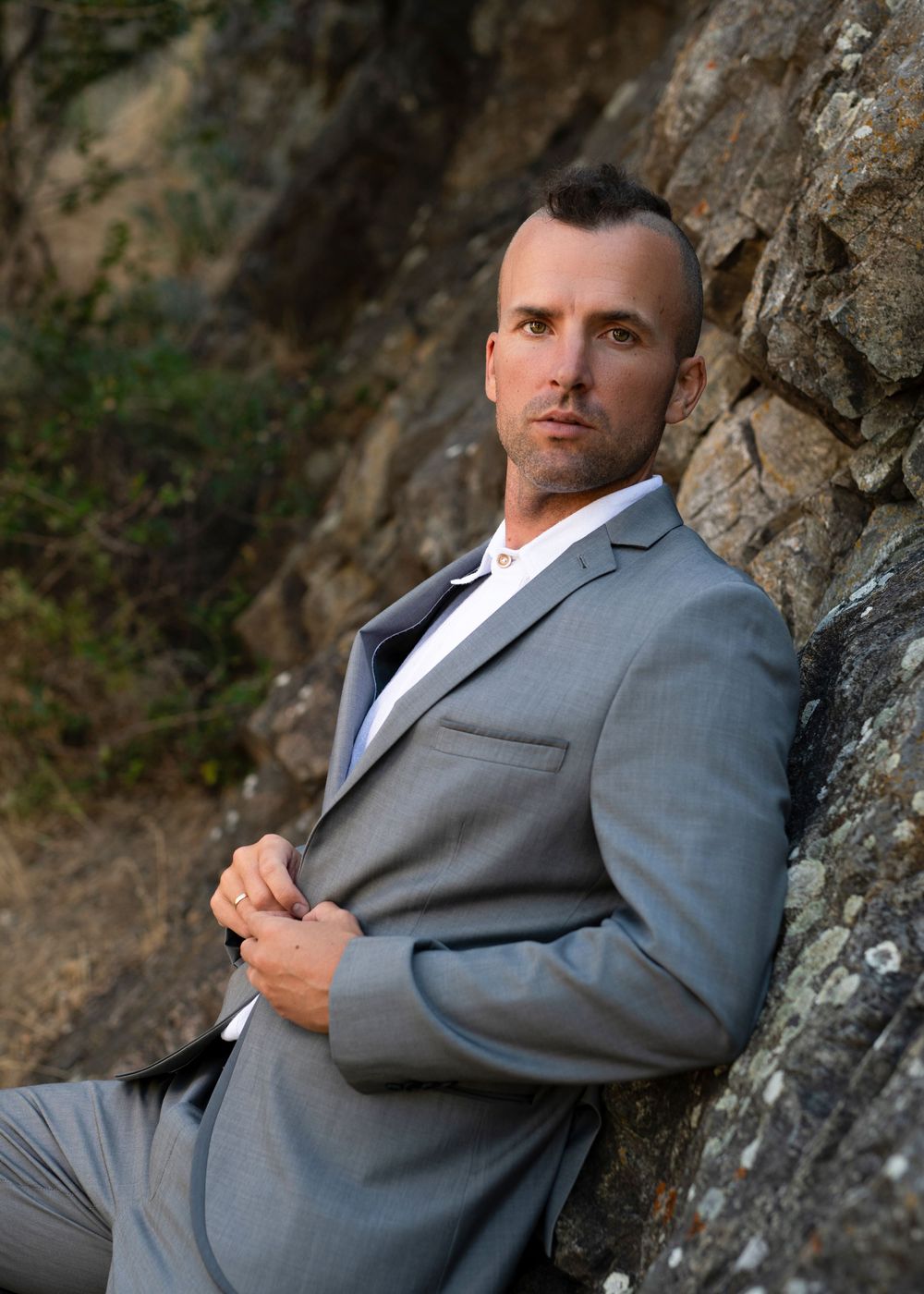 Young man in grey suit and white shirt buttoning jacket while leaning against a rock wall - Okanagan portrait by Tania Veitch.