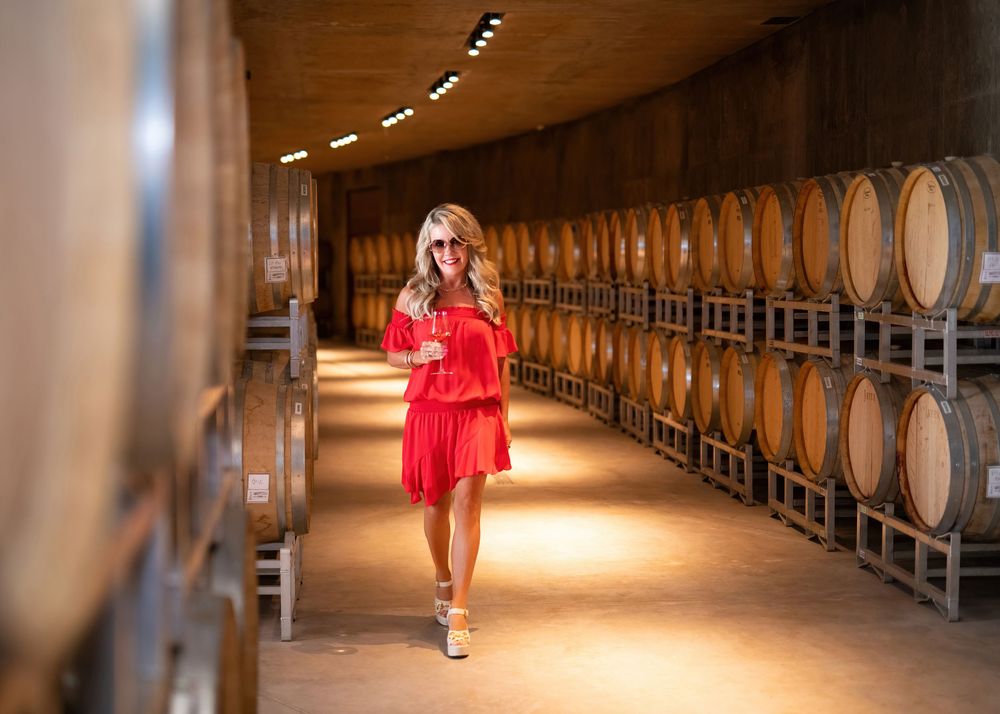 Founder of 50th Parallel Estate Winery in red dress and sunglasses walking through barrel cellar with glass of wine - photo by Tania Veitch.
