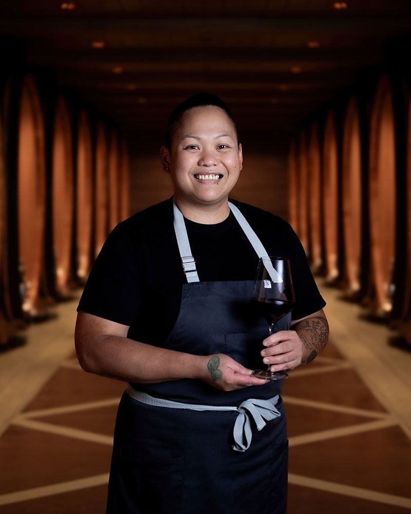 Former Phantom Creek Estates chef, Asian woman in apron and black shirt holding wine in barrel cellar - Okanagan portrait by Tania Veitch.