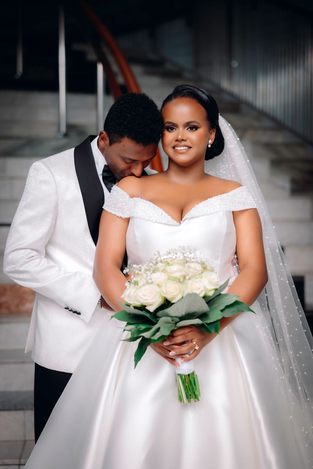 Elegant wedding couple in white attire sharing an intimate moment while holding a bouquet of white roses.