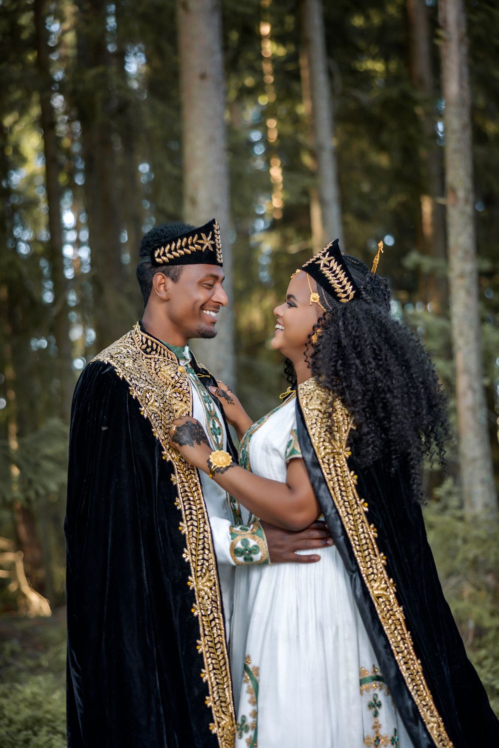 Wedding couple wearing matching black velvet capes with gold trim standing in a sunlit forest.