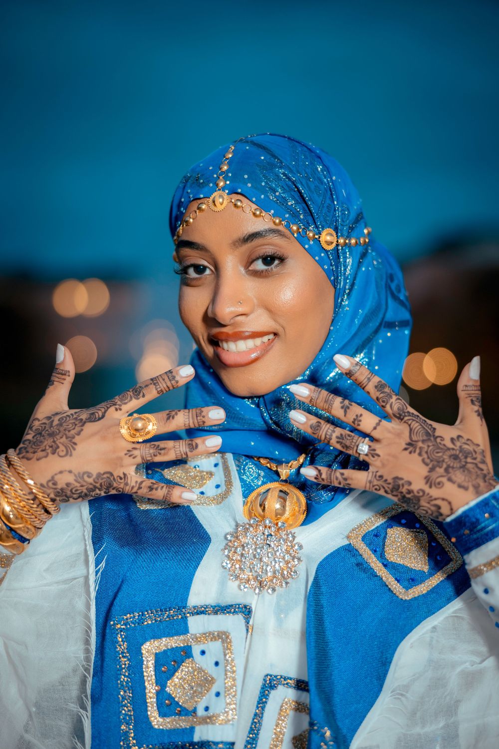 Woman in blue hijab with golden details displays henna-decorated hands and jewelry.