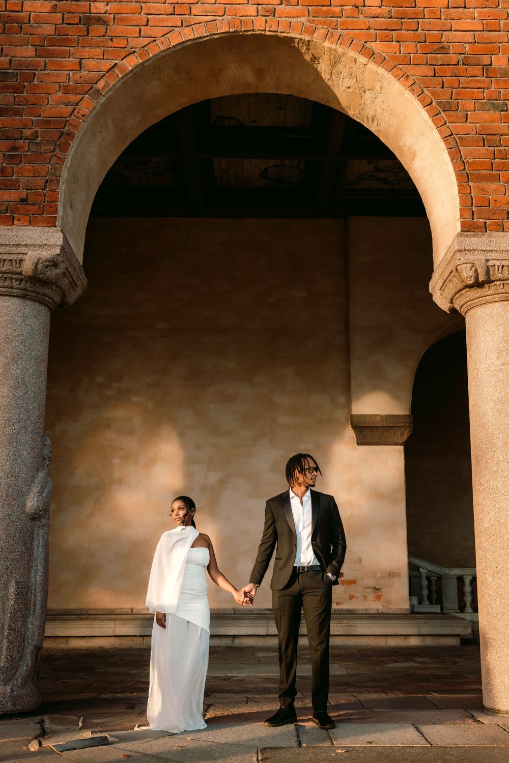 Couple in wedding attire holding hands under a stunning brick archway at sunset.