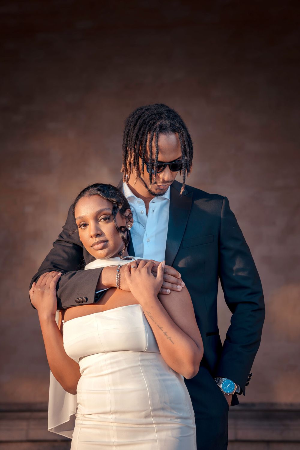 Stylish couple in formal attire posing for an intimate studio portrait against brown background.