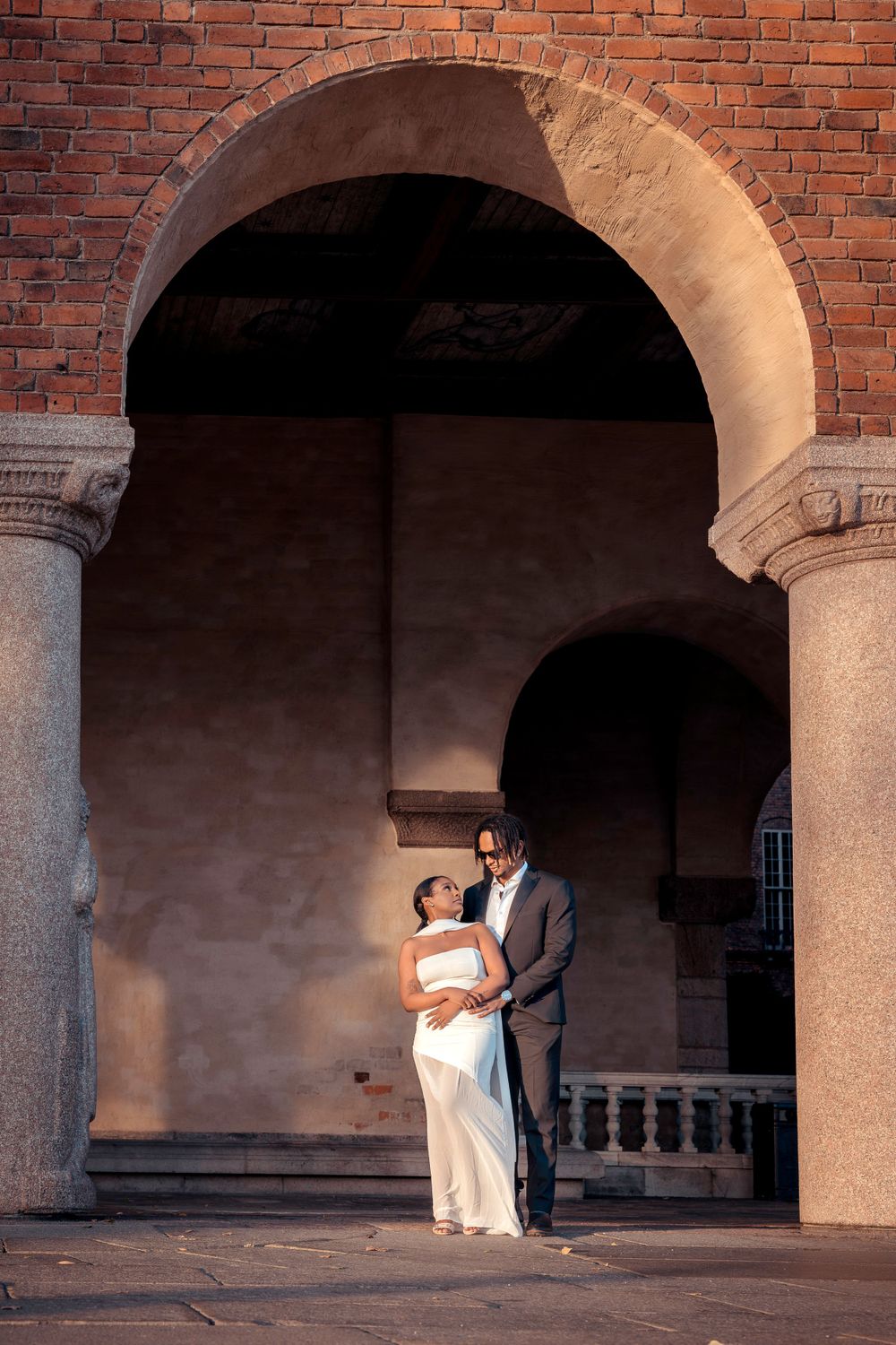 Wedding couple sharing tender moment under historic brick archway with dramatic lighting.