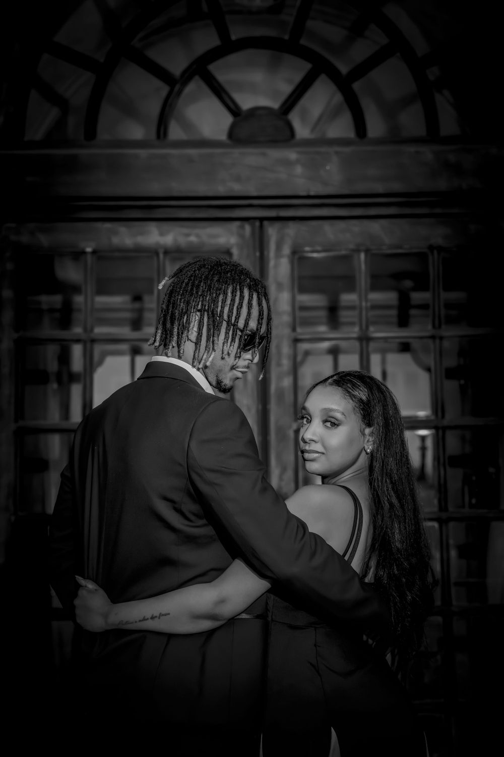 Romantic black and white portrait of couple embracing in front of ornate window architecture.