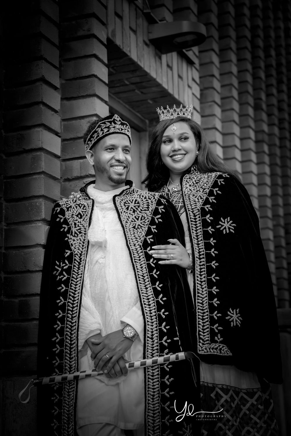 Couple wearing ornate black and gold royal-style robes and crowns pose against brick wall.