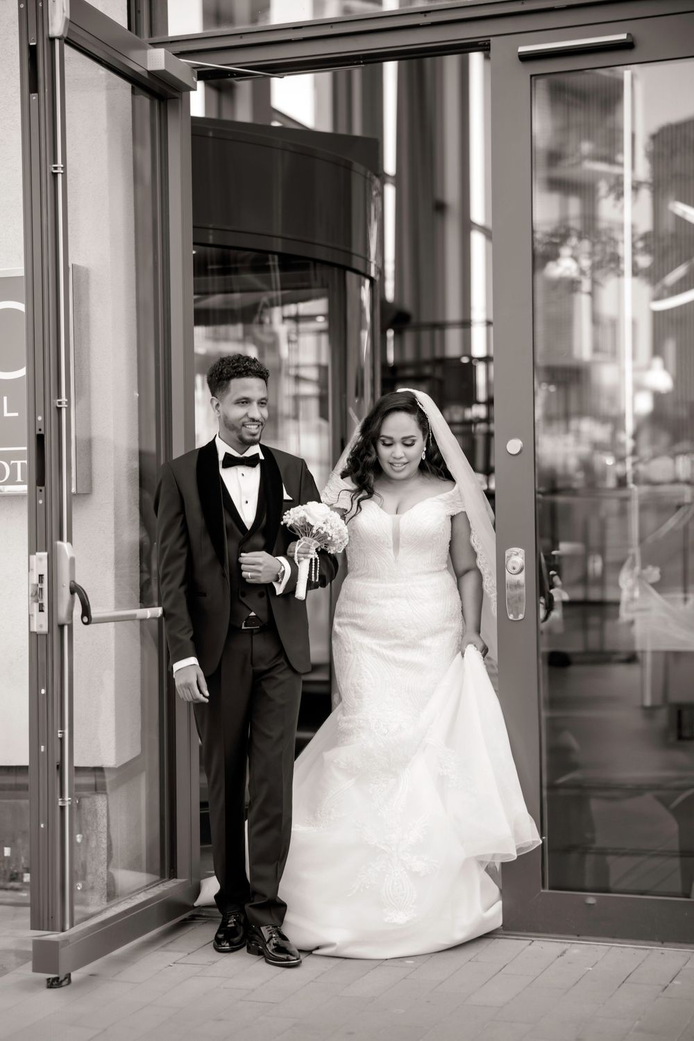 Black and white photo of newlyweds exiting modern glass building together.