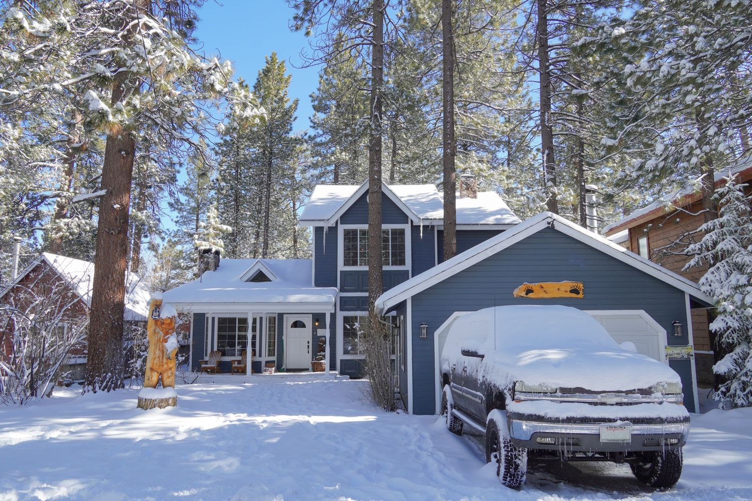 Snow-covered blue two-story home with attached garage surrounded by tall pine trees in a winter mountain setting.