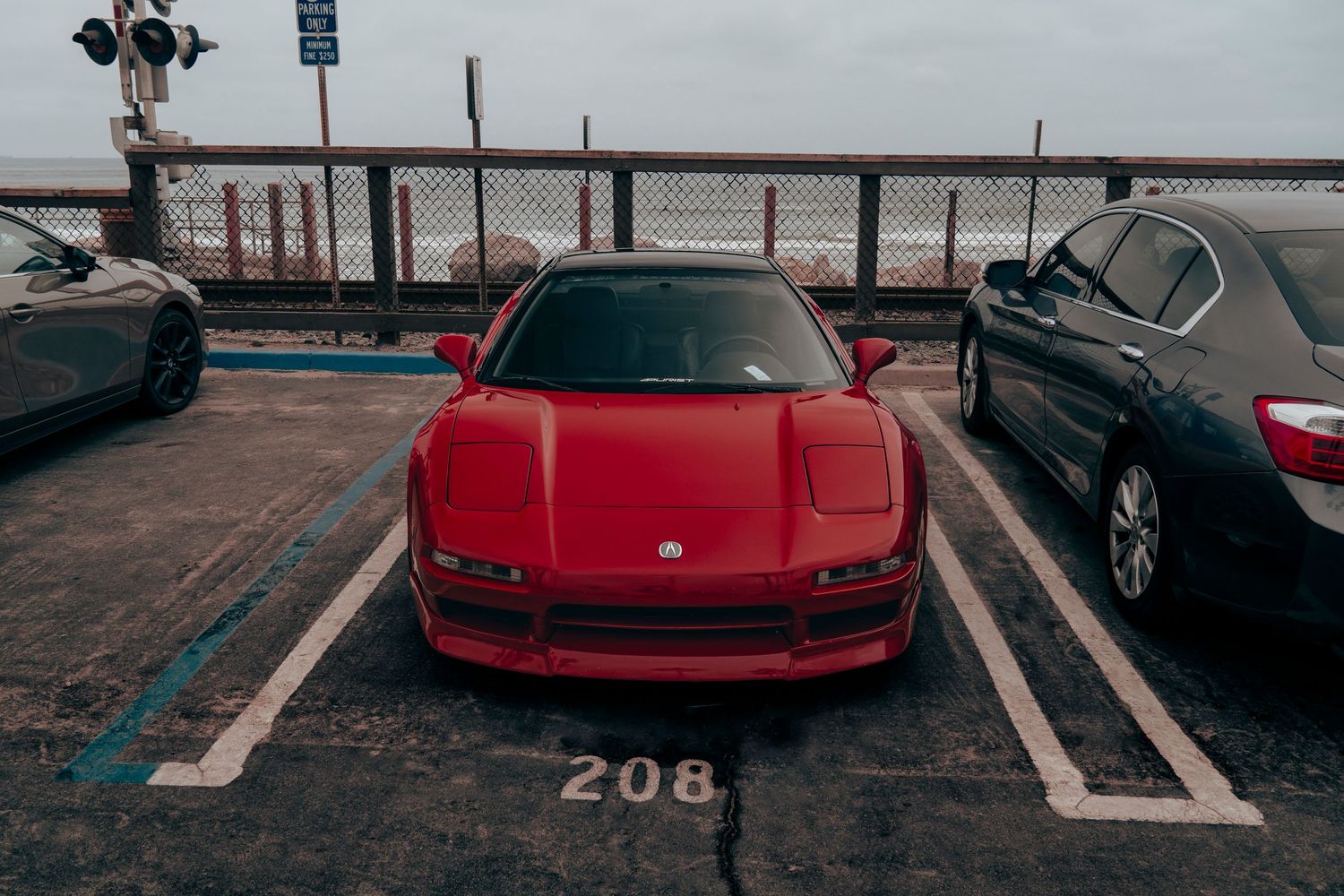 Red Acura NSX parked in spot 208 of a coastal parking lot on an overcast day.