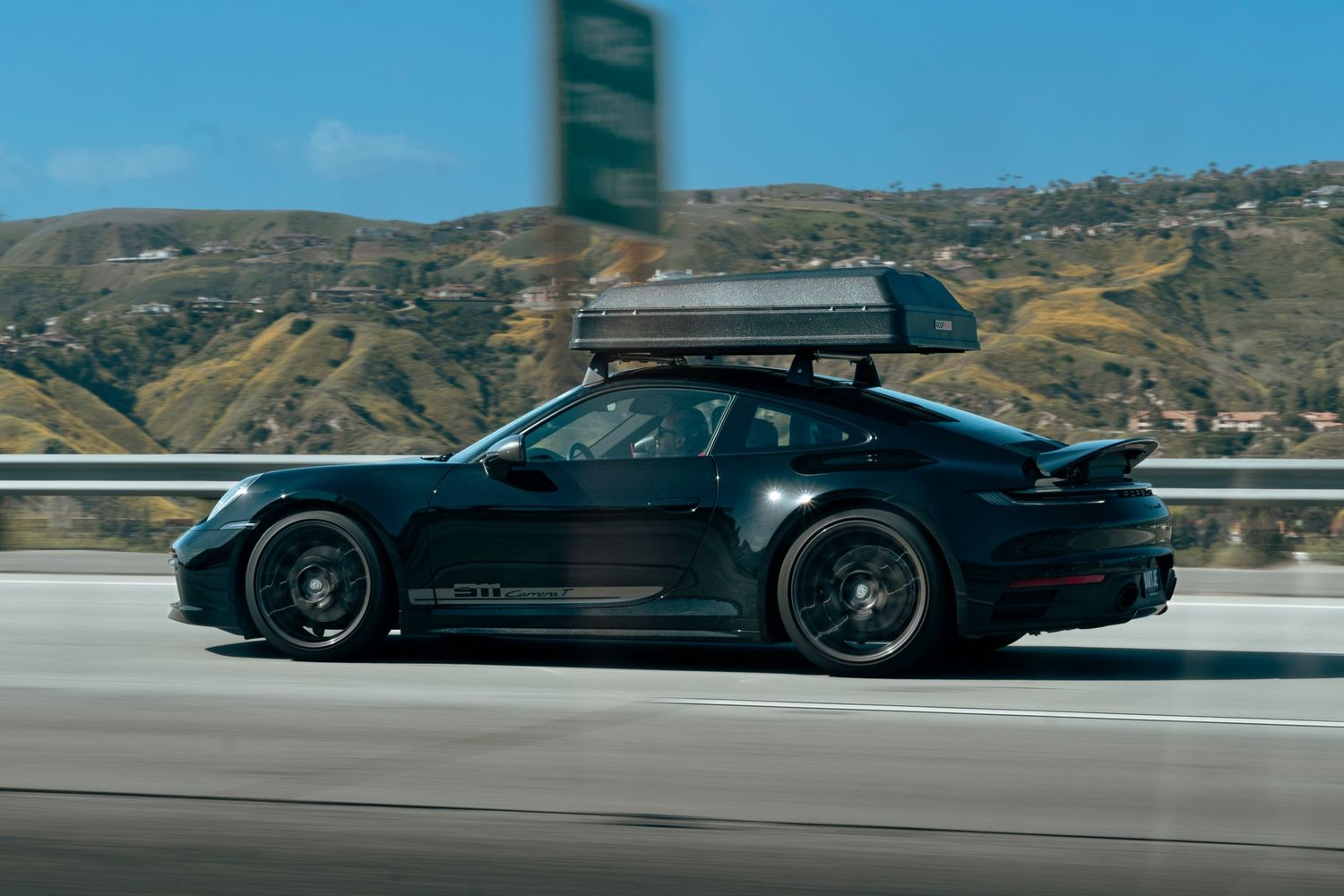 Dark green Porsche 911 GT3 with roof box driving on a California highway.