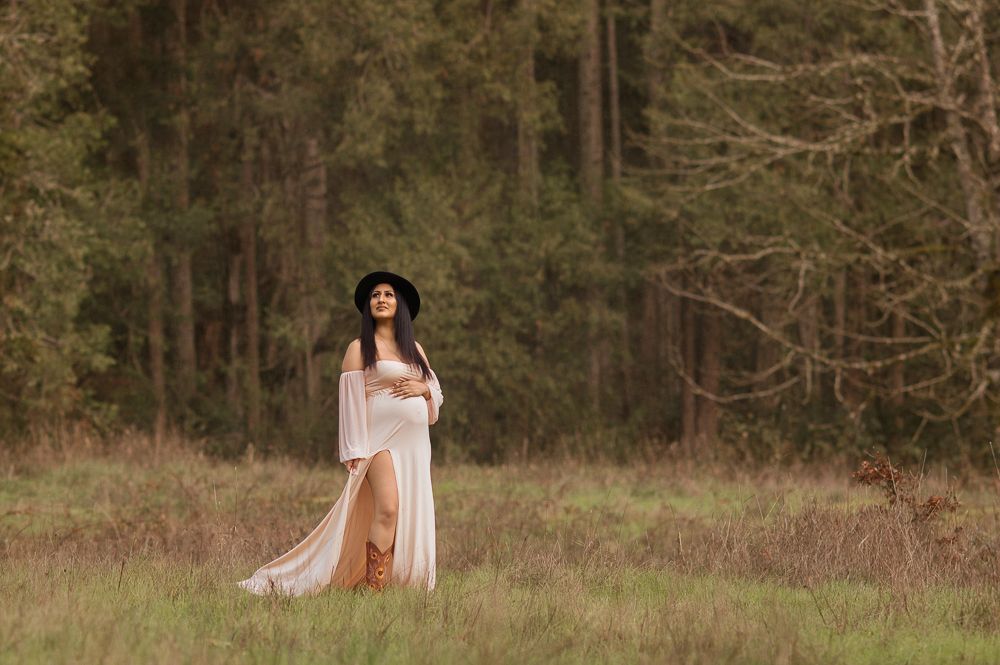 Woman in white boho dress and black hat standing in a grassy field with pine trees in the background.