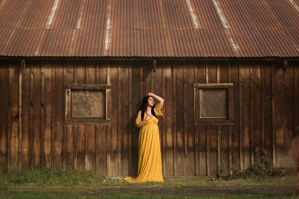 Woman in yellow maxi dress posing by rustic wooden barn with corrugated metal roof.