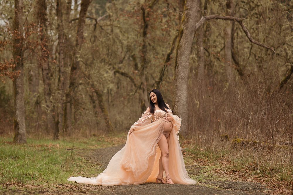 Woman in elegant peach gown posing gracefully on a forest path surrounded by bare trees in autumn.