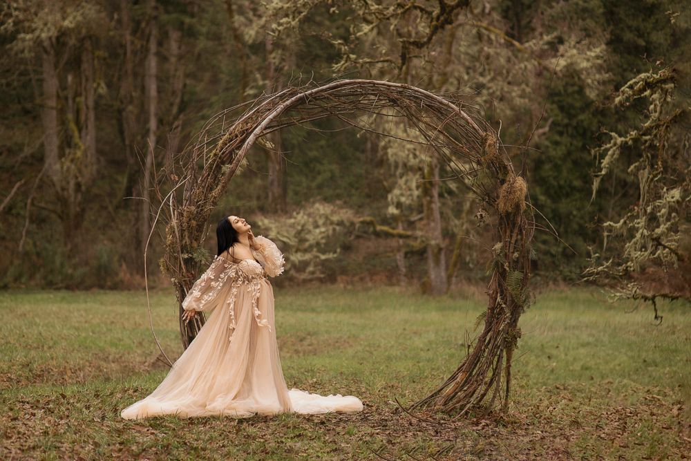 Woman in flowing gown posing under rustic twig arch in misty forest clearing.