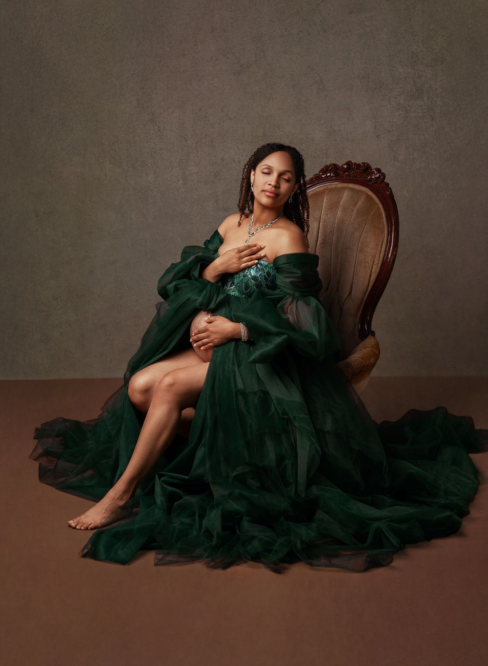 A person in an elegant emerald green tulle gown reclines gracefully on a vintage wooden chair against a neutral backdrop.