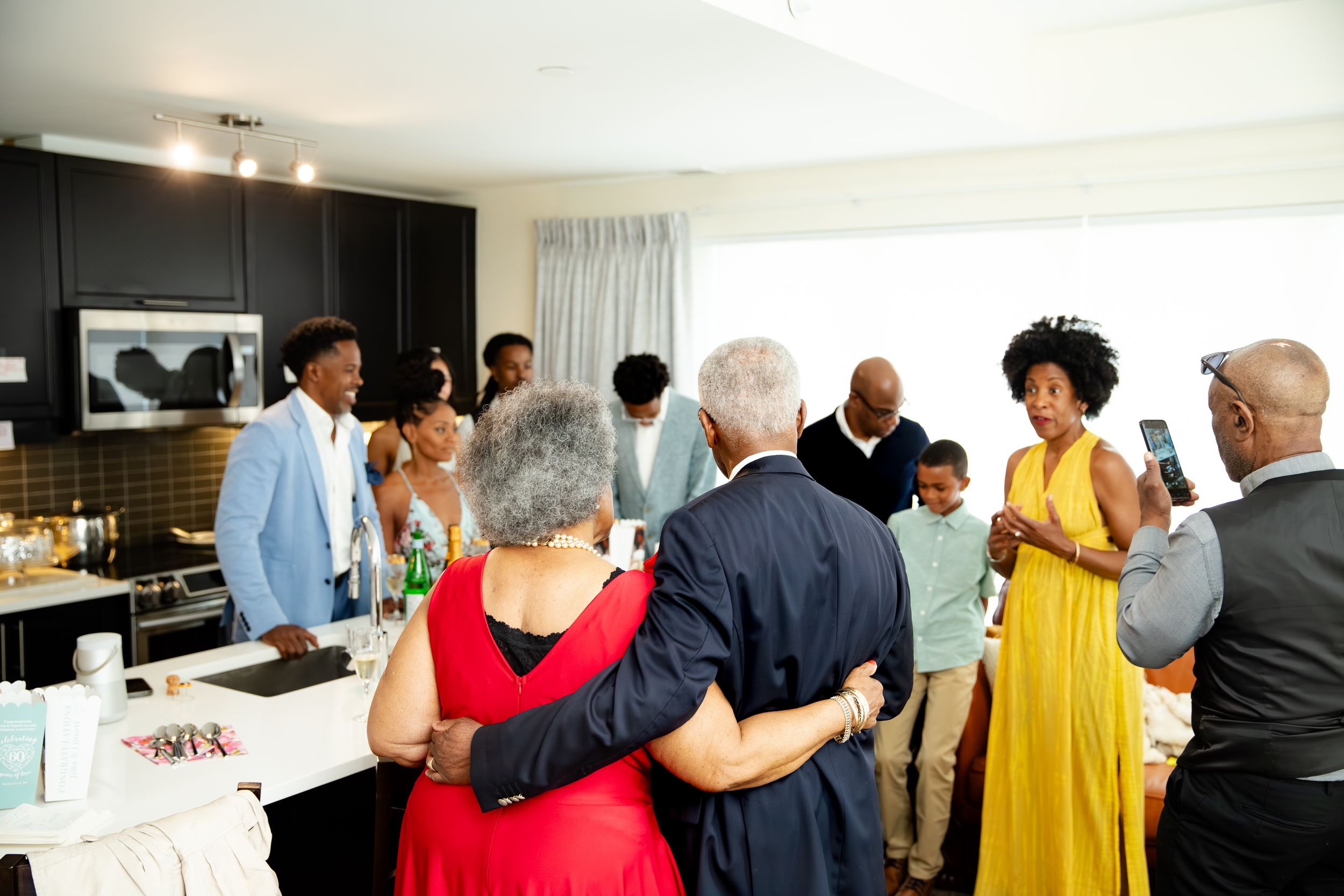 A diverse group of people gather in a modern kitchen space at a social event, with someone in a bright yellow dress standing out.