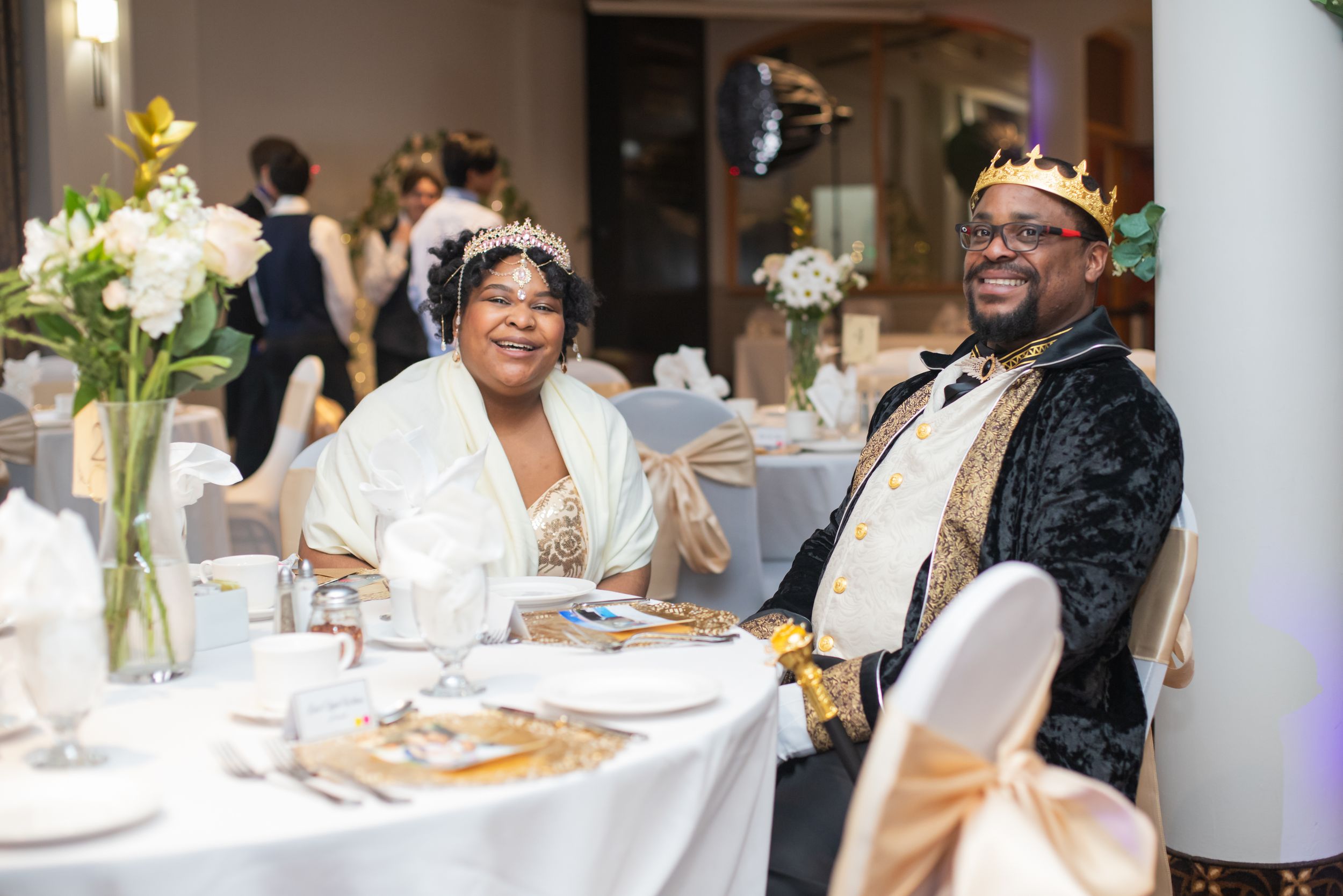 Two wedding guests sit at an elegantly decorated reception table with white floral centerpieces and formal place settings.