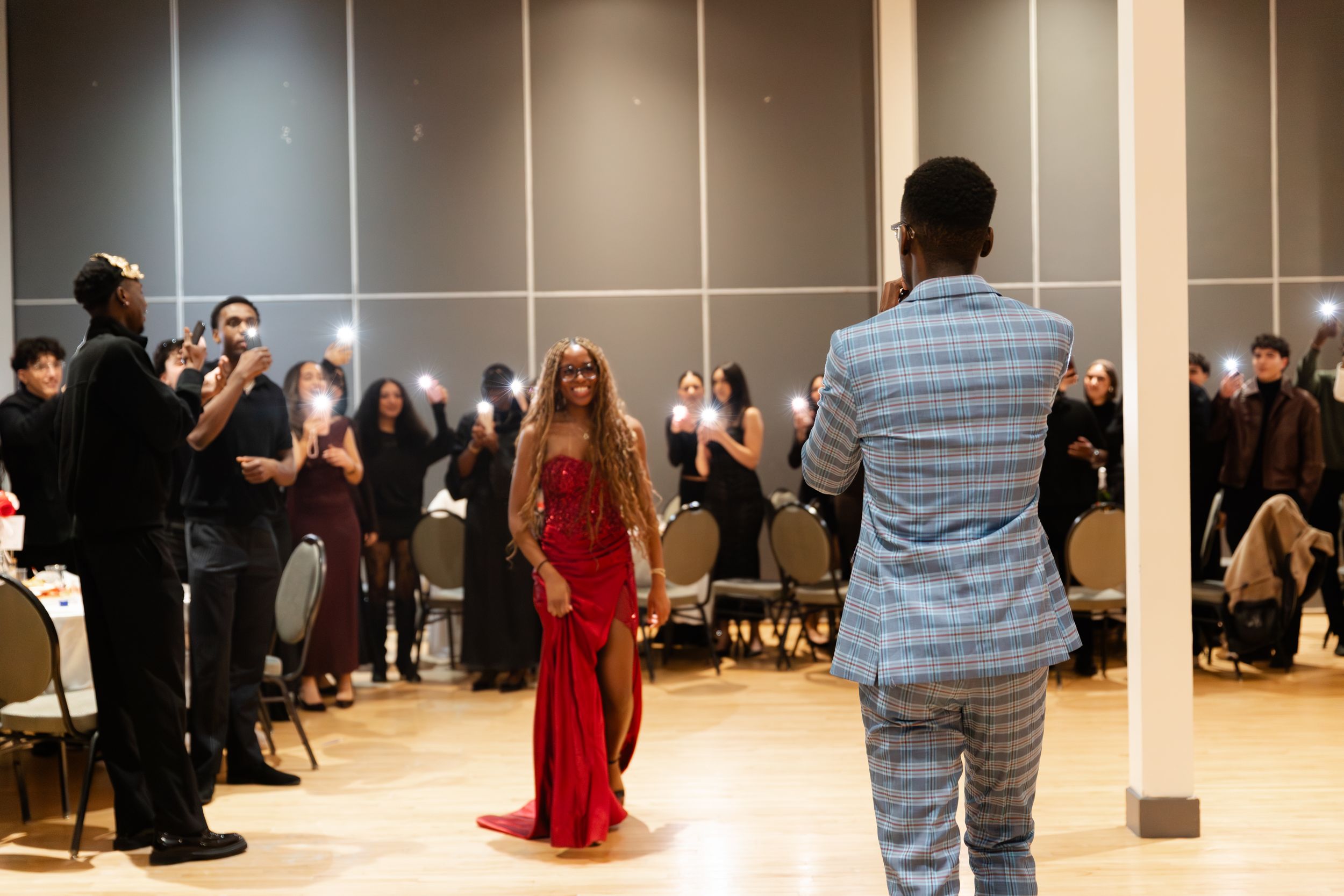 People gather in a performance space as someone in a red gown stands before someone wearing a blue striped suit.