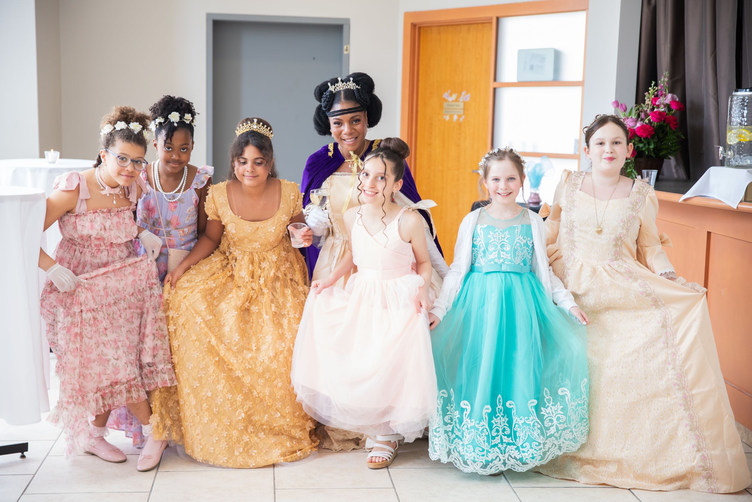 A group in elegant formal dresses and gowns pose together in a bright indoor setting.