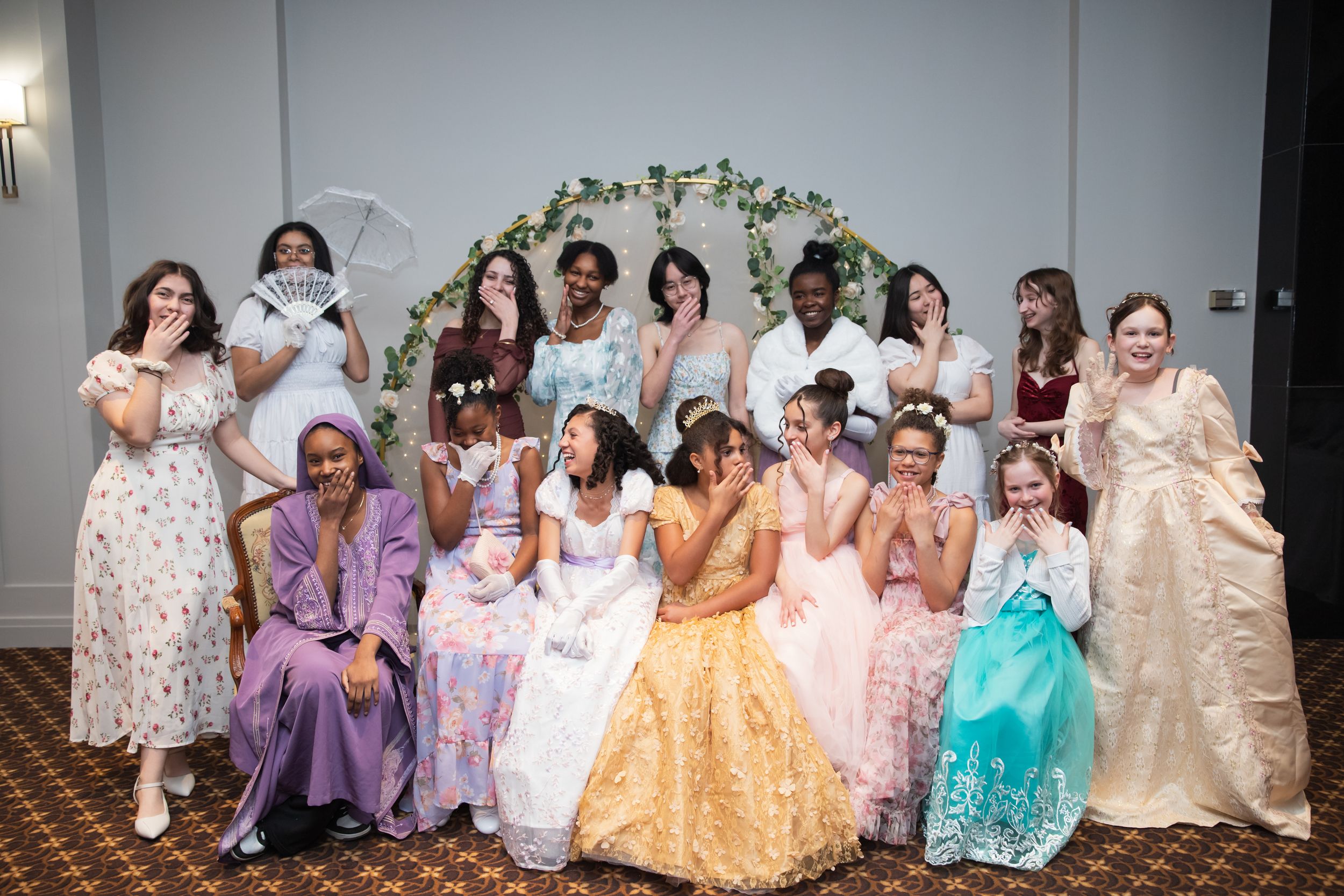 A group photo of wedding guests in pastel dresses sitting together on stairs for a formal portrait.