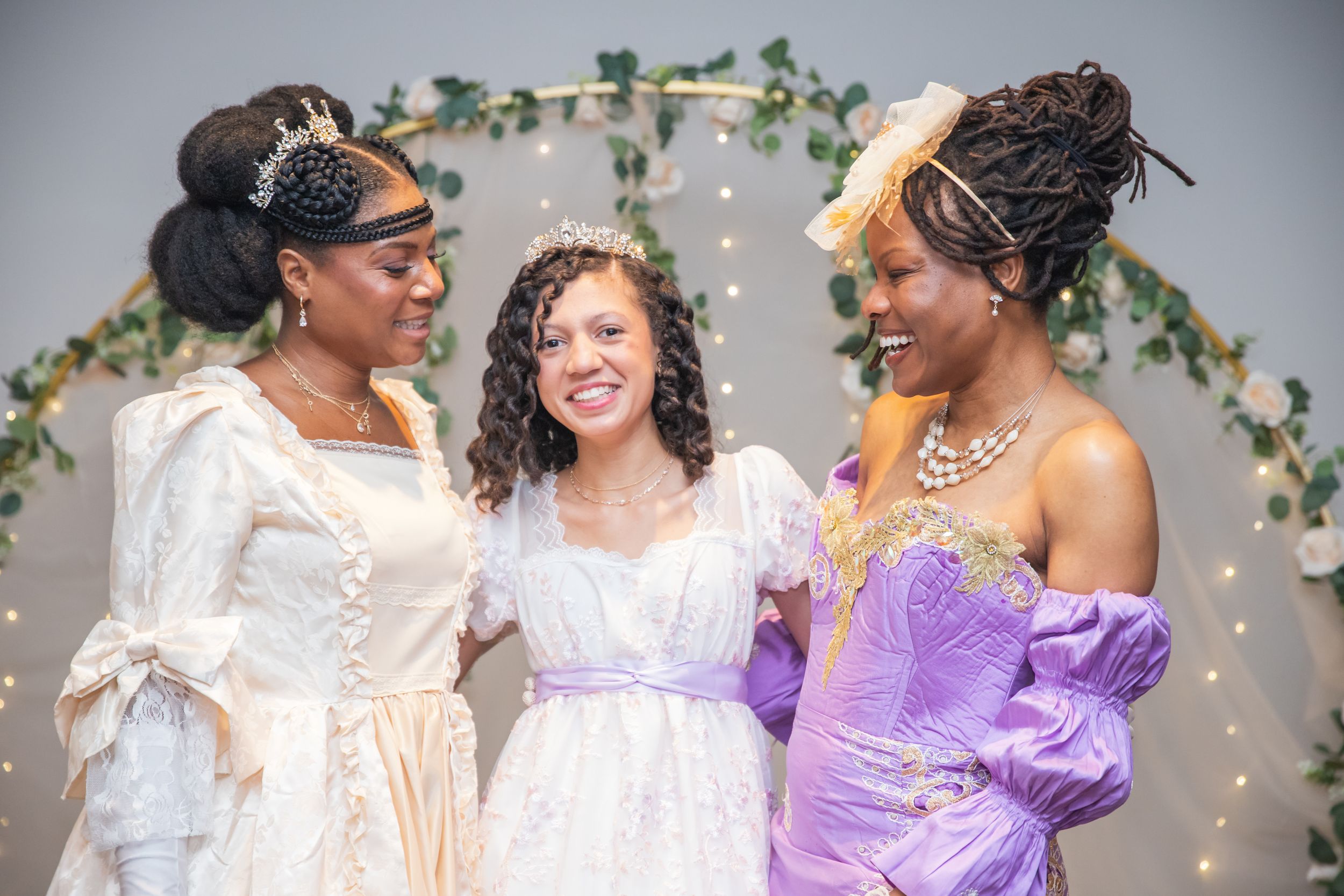 Three friends in elegant formal dresses laugh together in front of a floral arch with twinkling lights.