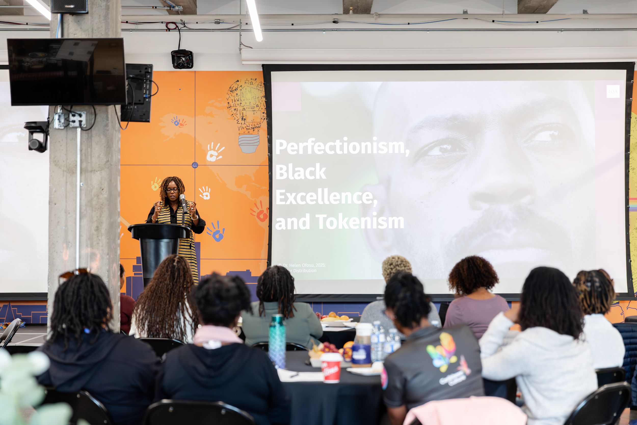 A speaker presents to an audience in a modern classroom with a projection screen displaying text about excellence.