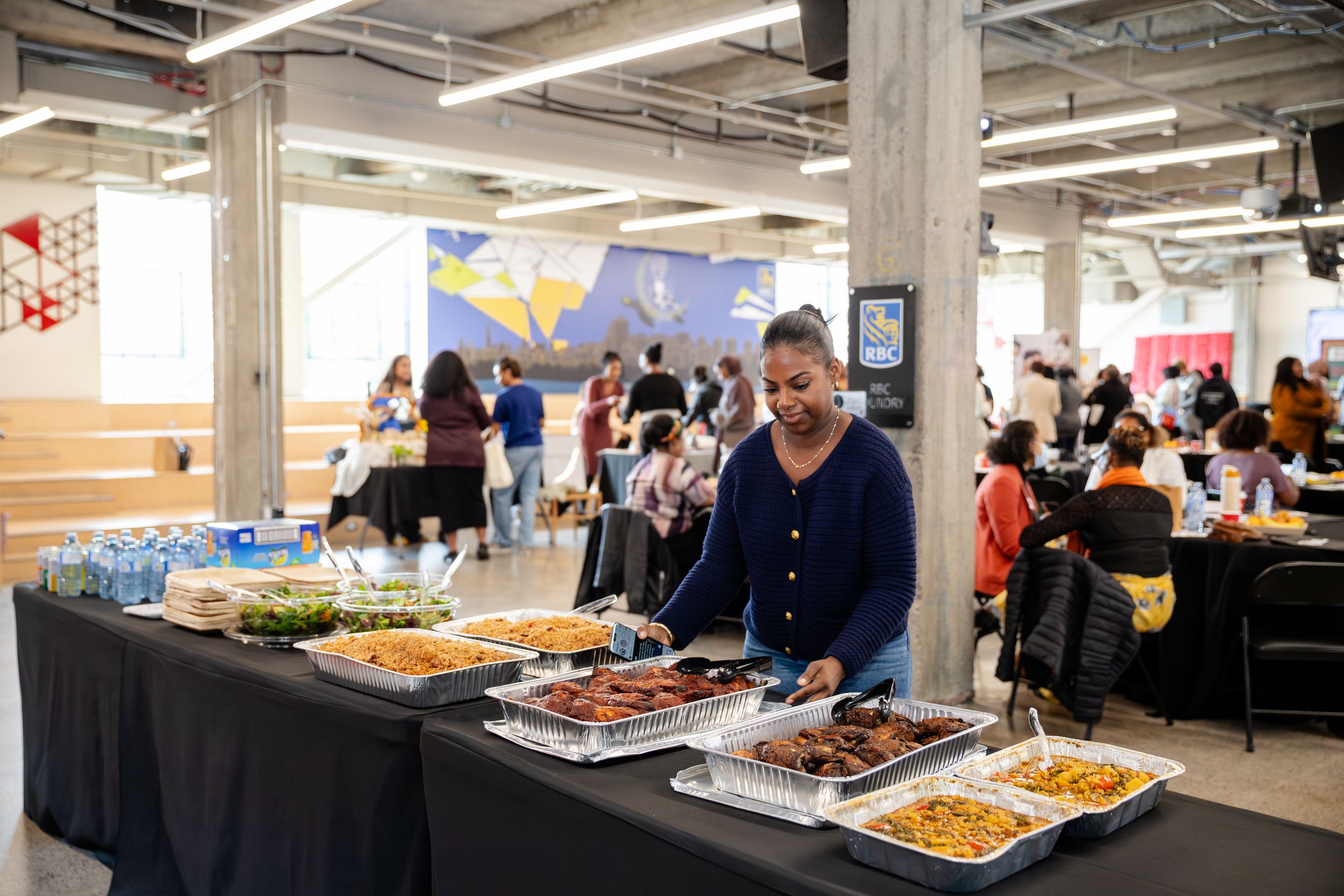 People gather around food stations at a cafeteria buffet with multiple serving trays displaying various dishes.