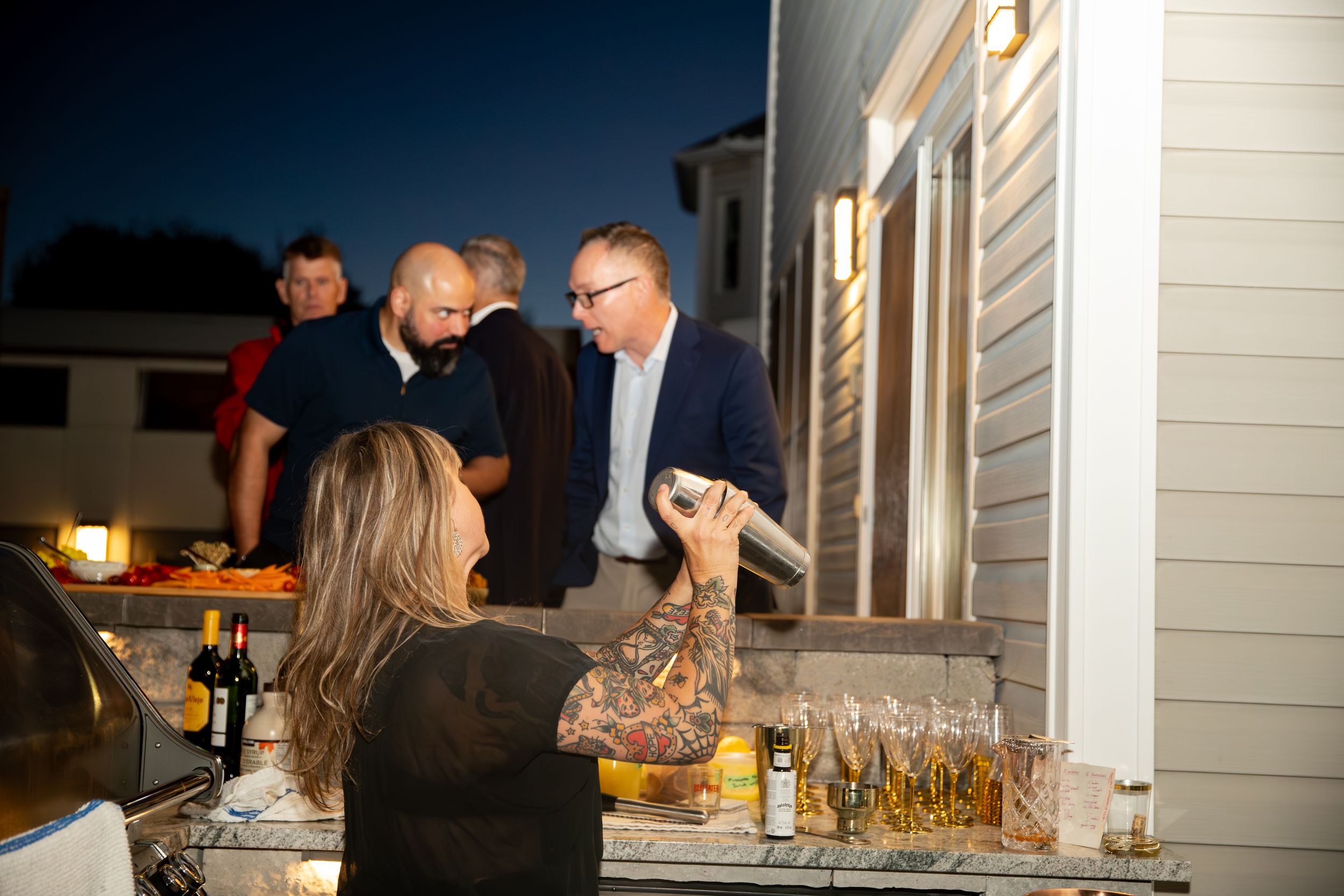 People gather on a patio at night for an outdoor dinner party with drinks and food spread across tables.
