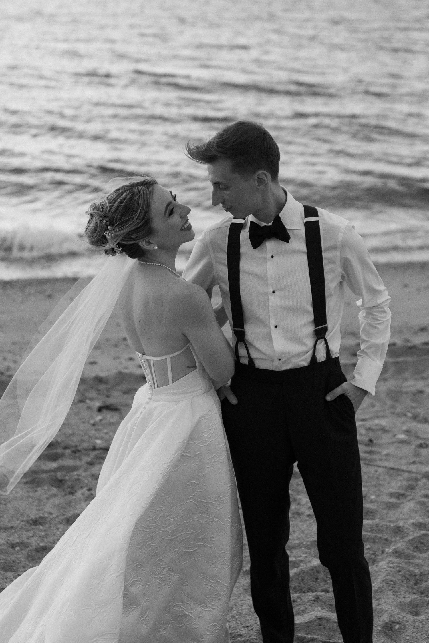 Romantic beach wedding photo session with flowing veil and formal attire.