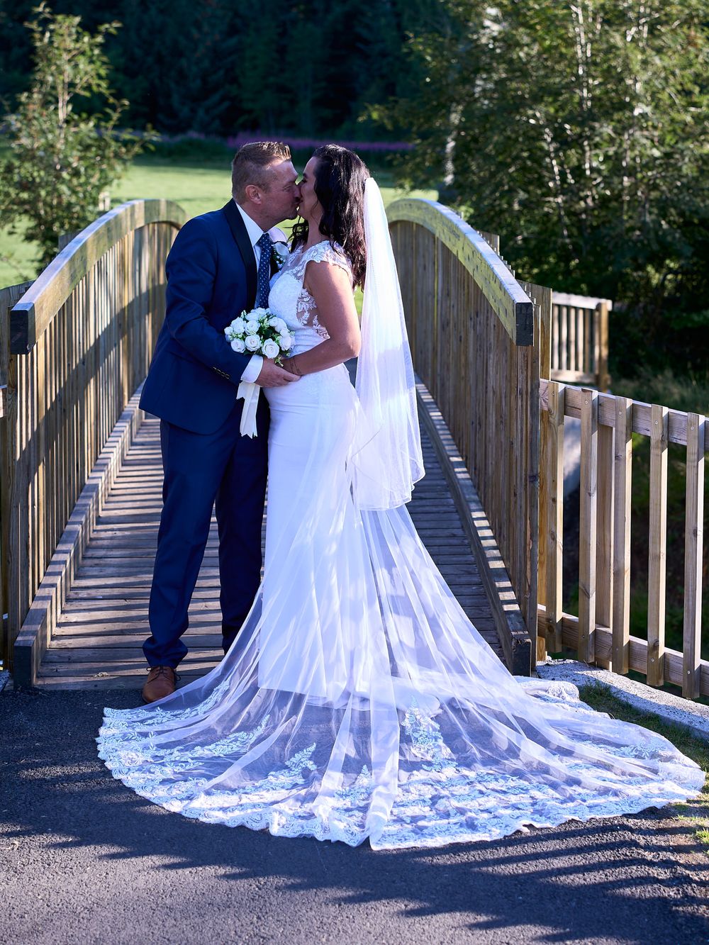 A romantic wedding couple shares a kiss on a wooden bridge as the bride's long lace train elegantly trails behind her.
