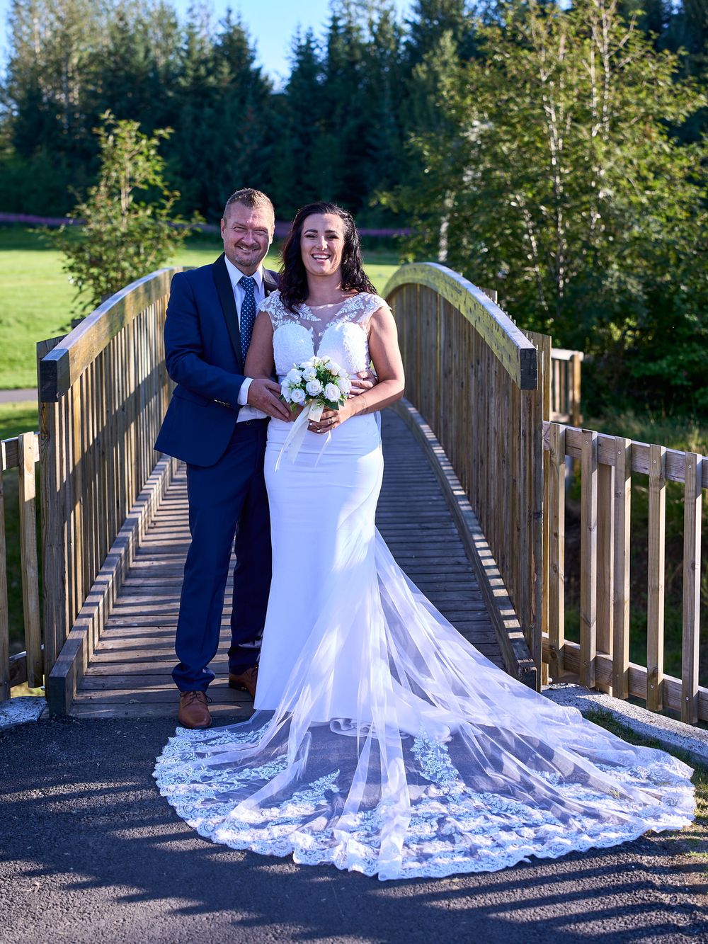 A wedding couple poses on a wooden footbridge surrounded by evergreen trees and sunlit foliage.