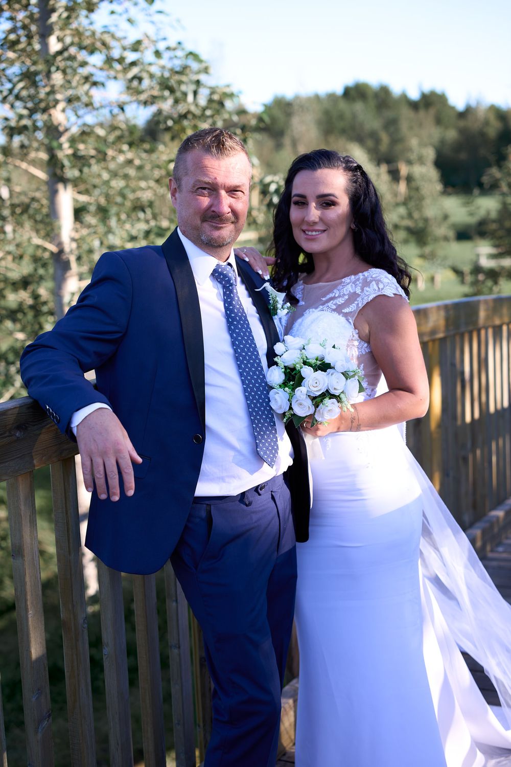 A couple poses together on a wooden deck at an outdoor wedding venue with lush greenery in the background.