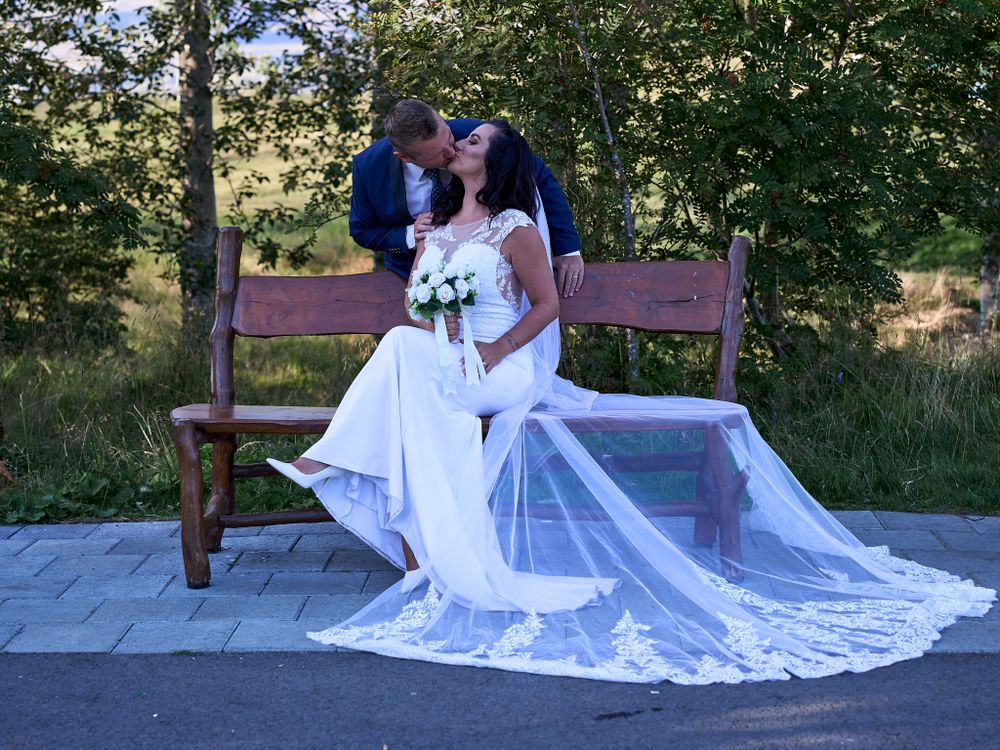 A couple in wedding attire sits on a wooden bench with a flowing white lace train spread out behind them.