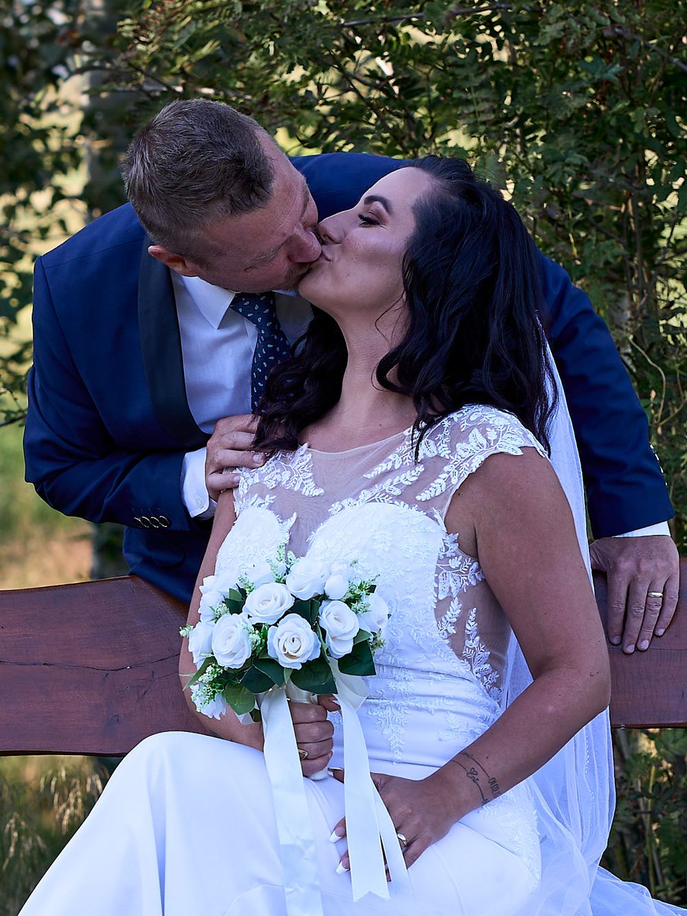 A romantic wedding couple shares a tender kiss while seated outdoors, with the bride holding a white rose bouquet.
